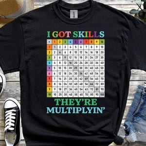 May include: Black t-shirt with a multiplication table graphic. The shirt features the text "I GOT SKILLS" at the top and "THEY'RE MULTIPLYIN'" at the bottom. A multiplication chart is in the centre, with numbers from 1 to 10.