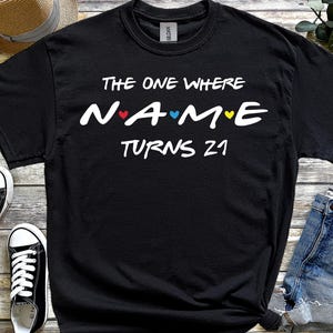 May include: Black t-shirt with white text that reads "THE ONE WHERE NAME TURNS 21". The word "NAME" has a red heart, a blue heart, and a yellow heart. The shirt is next to black and white trainers and denim shorts.