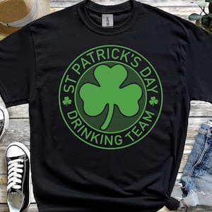 May include: Black t-shirt featuring a green shamrock and the words "ST PATRICK'S DAY DRINKING TEAM" in a circle. The shirt is styled with denim shorts and black and white trainers.