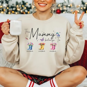 Puede incluir: Sudadera beige con la frase "This Mummy belongs To" y tres pares de botas de agua azules, amarillas y rosas, cada una con un nombre debajo. El diseño incluye mariposas y estrellas.
