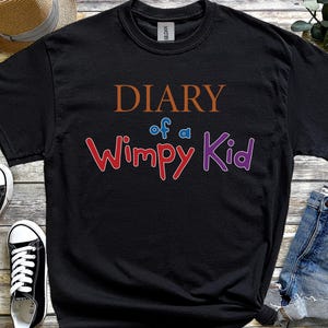 May include: Black t-shirt featuring the text "DIARY of a Wimpy Kid" in brown, blue, red, and purple. The shirt is shown with black and white trainers and denim shorts.