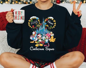 May include: A navy blue sweatshirt featuring a colorful graphic of Disney characters in front of a castle, with the text "Christmas Squad" and "2025". A mug with "Merry Christmas" is held in the hand.