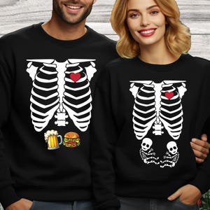 May include: Two black crewneck sweatshirts featuring a white ribcage graphic on the front. One sweatshirt displays a red heart, a beer mug, and a hamburger. The other sweatshirt features a red heart and two small skeleton figures at the bottom.