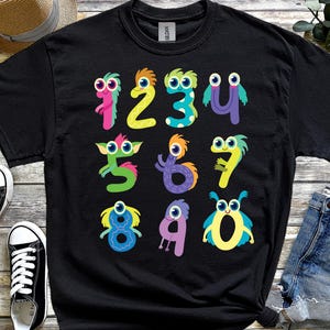 May include: Black t-shirt with cartoon numbers designed as monsters, each with googly eyes. The numbers 1 to 0 are in various bright colours. A pair of black and white trainers, a straw hat, and denim shorts are visible.