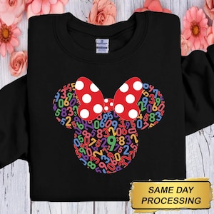 以下が含まれることがあります： ミニーマウスのデザインが施された黒いスウェットシャツ。デザインには、白い水玉模様の赤いリボンと、カラフルな数字で埋められたマウスの頭が含まれています。「SAME DAY PROCESSING」という言葉が金の長方形の中にあります。
