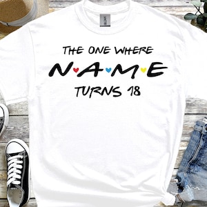 May include: White t-shirt with the text "THE ONE WHERE NAME TURNS 18" in black, with colourful hearts. The shirt is displayed with black and white trainers, denim shorts, and a straw hat.