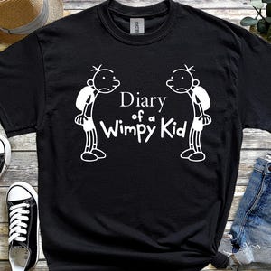 May include: Black t-shirt with the white text "Diary of a Wimpy Kid" and two cartoon figures. The image also shows a straw hat, black trainers, and denim shorts.