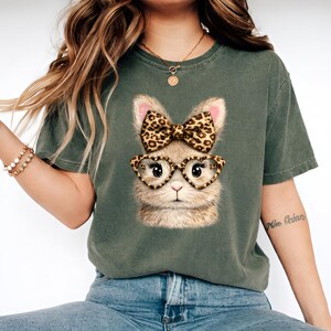 Puede incluir: Una camiseta de manga corta en color verde oliva oscuro con un estampado gráfico de una cara de conejo esponjoso. El conejo tiene orejas largas, un lazo con estampado de leopardo entre las orejas y gafas de corazón con estampado.