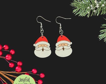 Op de afbeelding: Een paar kerst oorbellen met Santa Claus. Elke oorbel heeft een rode muts, een witte baard en een lachend gezicht. De oorbellen zijn van hout en hebben zilverkleurige haken. De afbeelding bevat de tekst "Joyful Laser Studio" en "SVG | Digital Download | Laser Tested".
