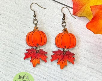 May include: A pair of fall-themed earrings featuring orange pumpkin pendants with brown stems and red and orange maple leaf charms. The earrings are suspended from bronze-colored ear wires. The image includes the text "Joyful Laser Studio" and "SVG | Digital Download | Laser Tested".