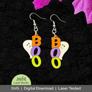 Könnte beinhalten: Ohrringe zum Thema Halloween mit dem Wort 'BOO'. Die Ohrringe zeigen einen Geist mit den Buchstaben B, O und O in Orange, Lila und Grün. Die Ohrringe sind aus Holz und haben silberne Haken.