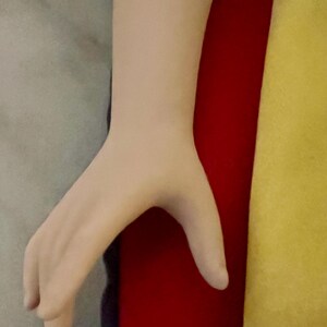 May include: A pale-toned hand with fingers slightly curved, positioned against a backdrop of gray, red, and yellow fabrics. The hand's skin appears smooth, with subtle shading.