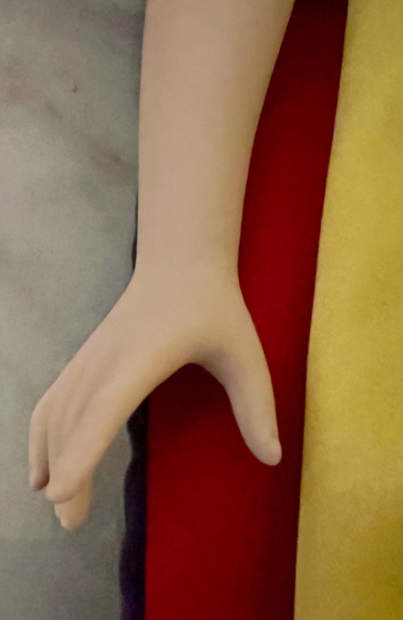 May include: A pale-toned hand with fingers slightly curved, positioned against a backdrop of gray, red, and yellow fabrics. The hand's skin appears smooth, with subtle shading.