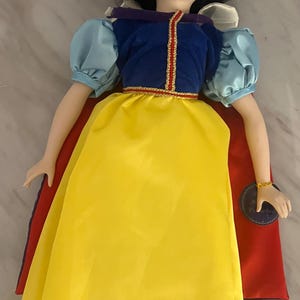 May include: A porcelain doll dressed in a yellow, blue, and red outfit. The doll has black hair, red lips, and a red bow. The doll's dress has a yellow skirt, a blue top with puffy sleeves, and a red cape.