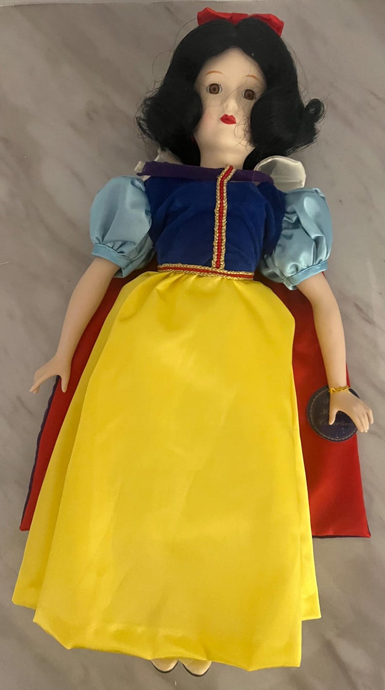 May include: A porcelain doll dressed in a yellow, blue, and red outfit. The doll has black hair, red lips, and a red bow. The doll's dress has a yellow skirt, a blue top with puffy sleeves, and a red cape.