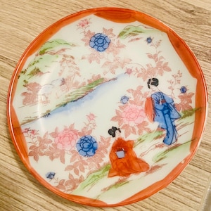 Könnte beinhalten: Ein runder, dekorativer Teller mit rotem Rand und weißem Innenraum. Der Teller zeigt eine detaillierte Illustration von zwei Figuren in traditioneller japanischer Kleidung, umgeben von floralen und landschaftlichen Elementen.