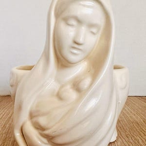 May include: Cream-colored ceramic planter in the shape of a woman with a draped head covering, holding a child. The figure has closed eyes and a serene expression. Two small openings are on either side of the head.
