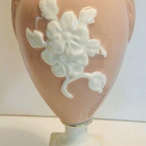 Puede incluir: Una lámpara de mesa con cuerpo de cerámica rosa pálido en forma de urna, con un relieve floral blanco en la parte frontal. La lámpara tiene dos asas laterales, una base cuadrada blanca y un borde dorado en la parte inferior de la base.