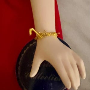May include: A porcelain doll's arm with a gold-colored bracelet. The arm is resting on a dark blue plate with a scalloped edge and the word "Collection" printed on it. The background is red.