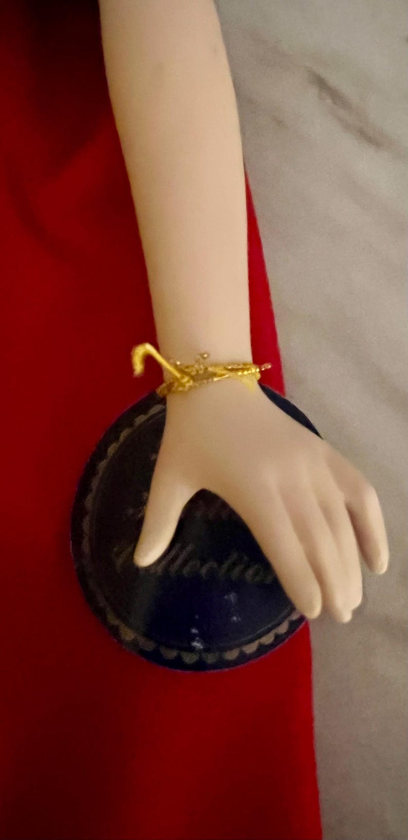 May include: A porcelain doll's arm with a gold-colored bracelet. The arm is resting on a dark blue plate with a scalloped edge and the word "Collection" printed on it. The background is red.
