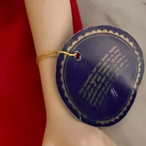 May include: A porcelain doll's arm with a gold bracelet and a blue tag. The tag has text that reads, "This exquisite doll has been made and the highest standards in the tradition of making..." The doll's arm is against a red background.