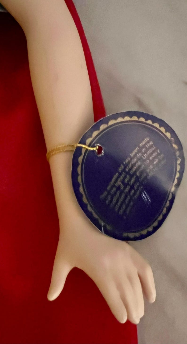 May include: A porcelain doll's arm with a gold bracelet and a blue tag. The tag has text that reads, "This exquisite doll has been made and the highest standards in the tradition of making..." The doll's arm is against a red background.