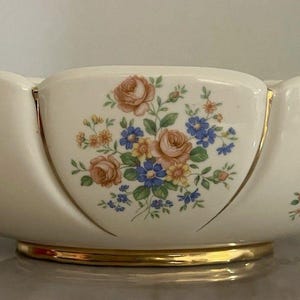 Puede incluir: Cuenco de cerámica color crema con borde festoneado y ribete dorado. El cuenco presenta ramos de flores en tonos rosa, azul y amarillo. Hay asas doradas en espiral en cada extremo. El cuenco se apoya sobre una base dorada.