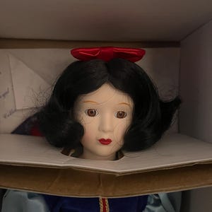 May include: A porcelain doll of Snow White, with black hair, a red bow, and a blue and yellow dress. The doll is in a cardboard box, likely a collector's item.