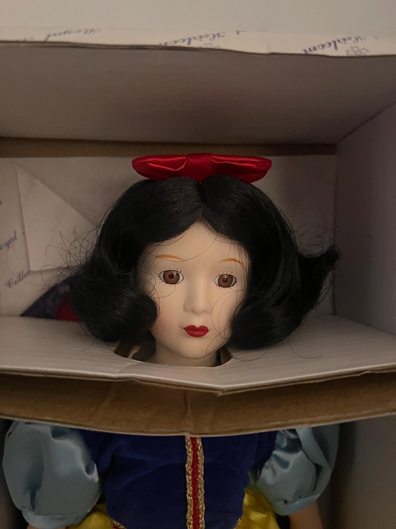 May include: A porcelain doll of Snow White, with black hair, a red bow, and a blue and yellow dress. The doll is in a cardboard box, likely a collector's item.