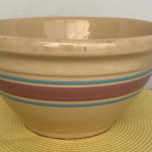 Puede incluir: Un cuenco mezclador de cerámica beige con un borde ancho y una forma redondeada. El cuenco presenta dos rayas horizontales: una ancha raya granate entre dos rayas turquesas más finas. El cuenco está sobre una superficie amarilla.