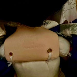 May include: A vintage doll with black hair and a tan plastic back. The back of the doll has the text "ANANDO" printed on it. The doll is wearing a white dress and a yellow skirt.