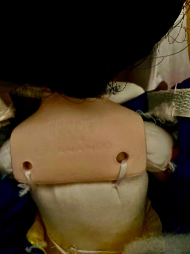 May include: A vintage doll with black hair and a tan plastic back. The back of the doll has the text "ANANDO" printed on it. The doll is wearing a white dress and a yellow skirt.
