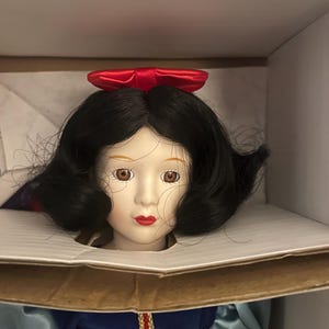 May include: A porcelain doll of a princess with black hair, brown eyes, and red lips. She wears a blue and yellow dress with a red bow in her hair. The doll is in a cardboard box.