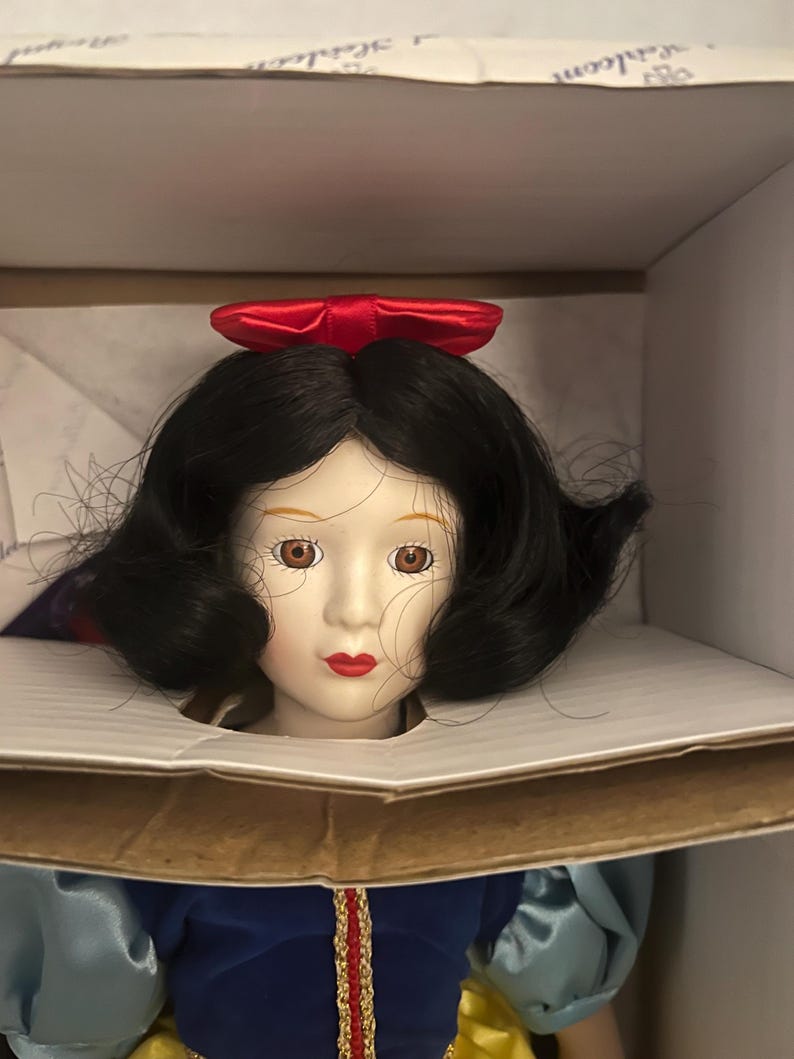 May include: A porcelain doll of a princess with black hair, brown eyes, and red lips. She wears a blue and yellow dress with a red bow in her hair. The doll is in a cardboard box.