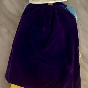 May include: A doll wearing a purple velvet cape and a yellow satin dress. The doll has black hair and a white collar. The doll's outfit is reminiscent of a fairytale character.