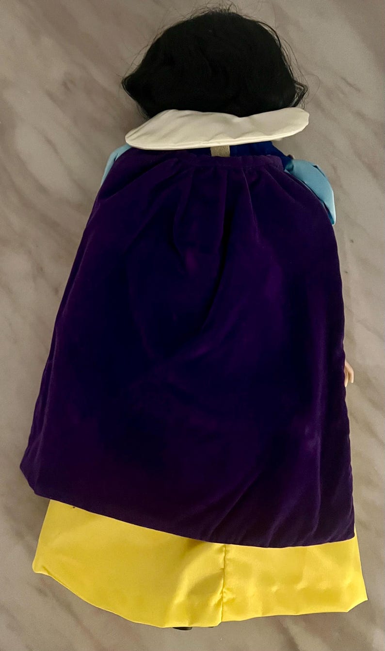 May include: A doll wearing a purple velvet cape and a yellow satin dress. The doll has black hair and a white collar. The doll's outfit is reminiscent of a fairytale character.