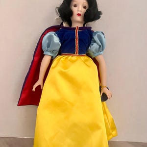 May include: A porcelain doll dressed as Snow White, featuring a yellow satin skirt, a blue velvet bodice, and a red cape. The doll has black hair, a red bow, and a detailed painted face. The doll is a collectible item.