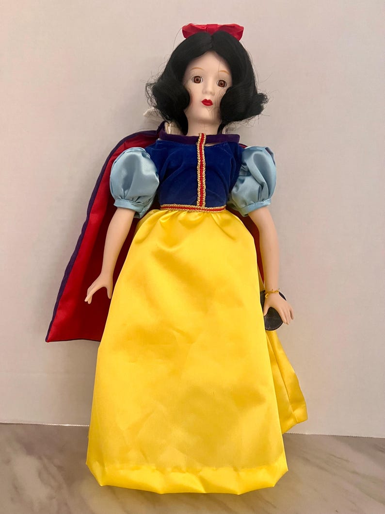 May include: A porcelain doll dressed as Snow White, featuring a yellow satin skirt, a blue velvet bodice, and a red cape. The doll has black hair, a red bow, and a detailed painted face. The doll is a collectible item.