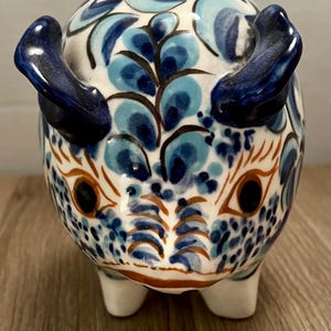 Puede incluir: Una alcancía de cerámica con un diseño detallado pintado a mano. El banco presenta una base blanca con patrones florales azules y detalles, incluyendo orejas y detalles faciales azul oscuro. La alcancía tiene una ranura en la parte superior para insertar monedas.
