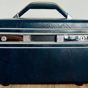 Puede incluir: Un neceser vintage azul marino con exterior texturizado, ribetes plateados y asa negra. La maleta tiene dos cierres plateados con cerraduras y una correa de cuero negro. La maleta es rectangular con esquinas redondeadas.