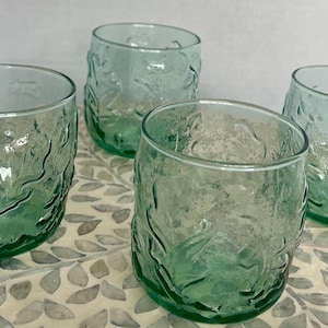 Puede incluir: Cuatro vasos transparentes teñidos de verde. Cada vaso tiene un patrón texturizado en forma de hoja en el exterior. Los vasos son cilíndricos con un borde ligeramente inclinado. Están dispuestos sobre una superficie estampada.