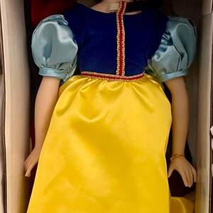May include: A porcelain doll of a princess in a box. The doll wears a blue velvet bodice with a gold trim, a yellow satin skirt, and puffy blue sleeves. A red bow adorns her black hair.