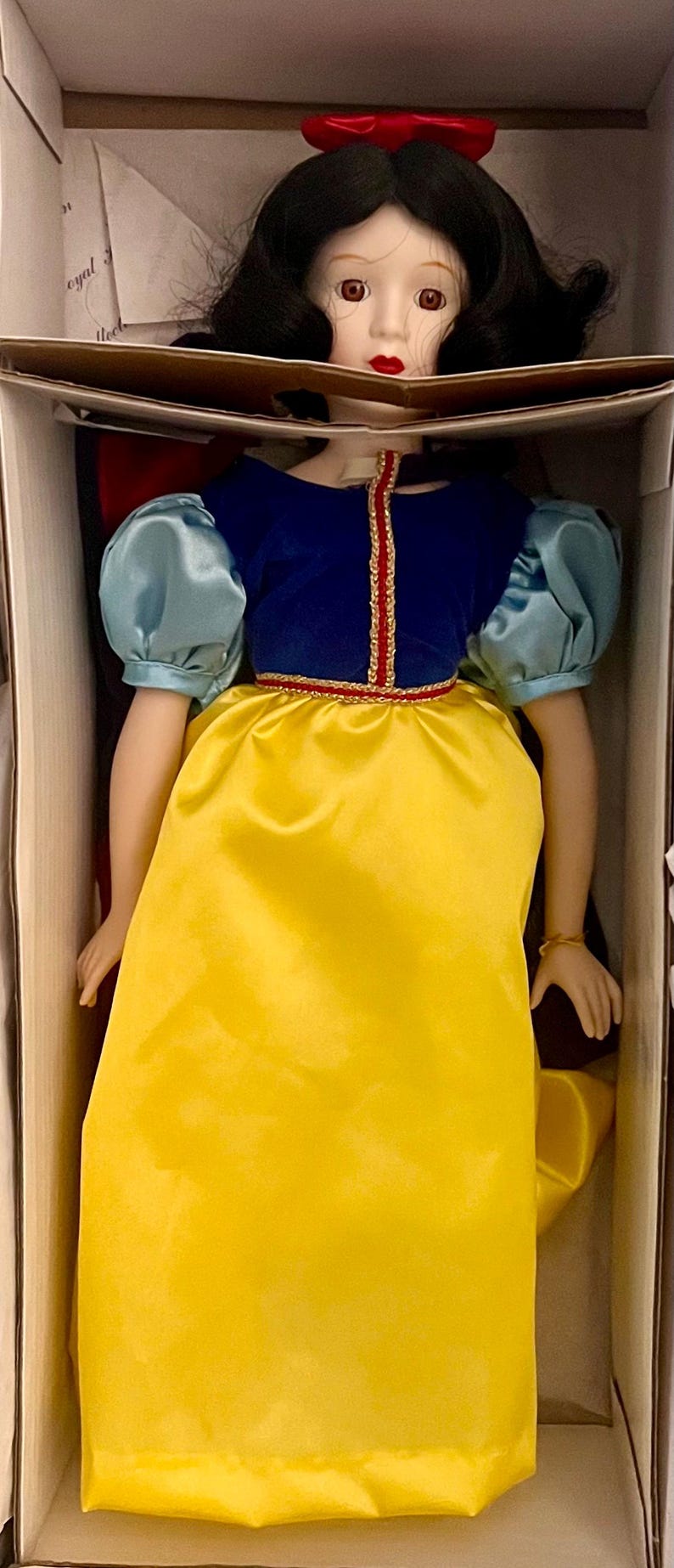 May include: A porcelain doll of a princess in a box. The doll wears a blue velvet bodice with a gold trim, a yellow satin skirt, and puffy blue sleeves. A red bow adorns her black hair.