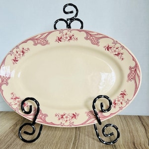 May include: An oval-shaped cream-colored ceramic platter featuring a decorative border of dark red floral patterns and intricate lace-like detailing, displayed on a black metal wire stand with scrolled feet and a top loop for support.