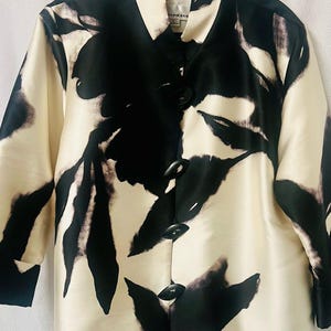 May include: Cream-colored jacket with a black floral pattern and black buttons. The jacket has a collared neckline and long sleeves. The floral design is abstract and covers the entire jacket. The jacket is buttoned up.