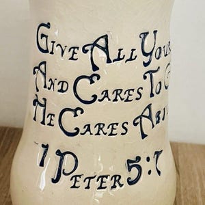 May include: Cream ceramic vase with a gold rim and dark blue lettering. The text reads "Give All Your Cares To Him, He Cares About You, 1 Peter 5:7". The vase has a bulbous base and a slightly flared top.