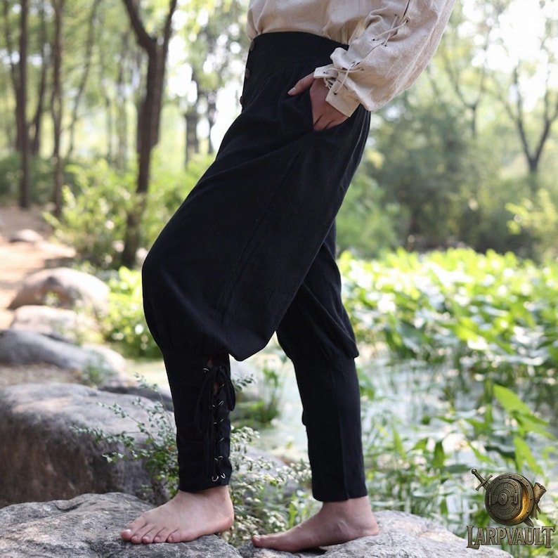 May include: Black medieval pants with a high waist and a loose fit around the thighs. The pants have a laced detail at the ankles and pockets. The person is standing on a rock in a natural setting.