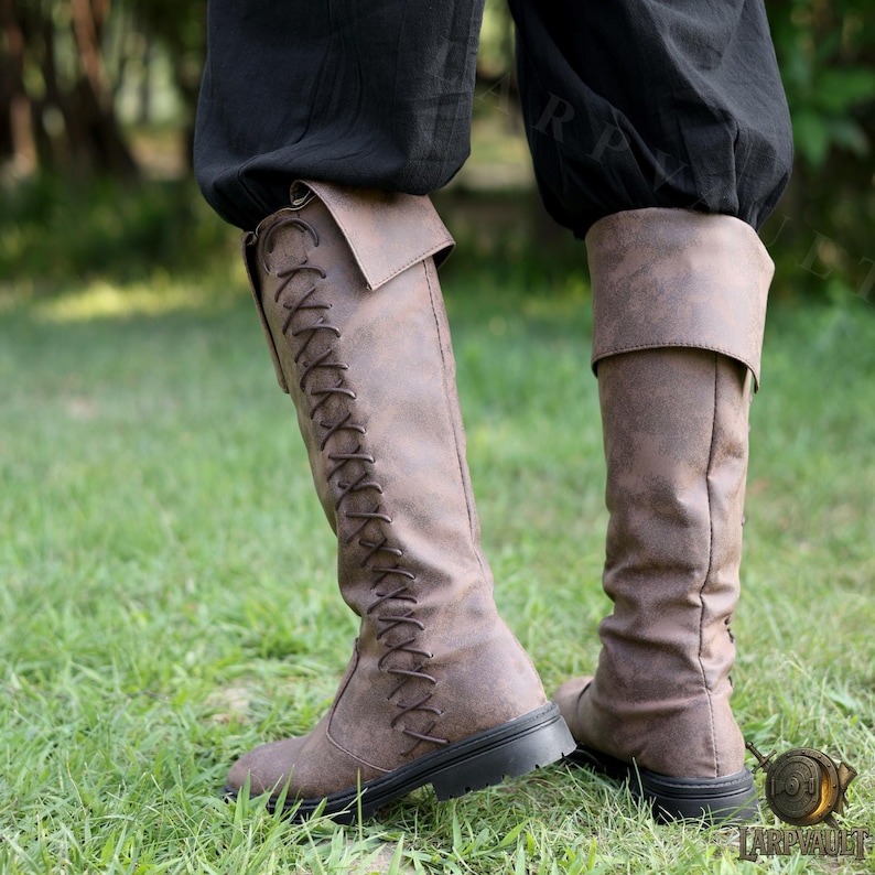 Puede incluir: Botas altas de cuero marr&oacute;n con dise&ntilde;o de cordones en la parte delantera. Las botas tienen una parte superior doblada y una suela negra. Las botas se usan con pantalones negros y est&aacute;n sobre hierba verde.
