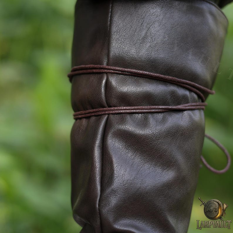 May include: Close-up of a brown leather boot with a textured surface. The boot is wrapped with a darker brown leather cord around the calf area. The background is a blurred green.