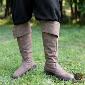 Puede incluir: Botas marrones hasta la rodilla con un pu&ntilde;o doblado. Las botas tienen un aspecto desgastado y texturizado y un tac&oacute;n bajo. Las botas se llevan con pantalones negros, sobre hierba verde.